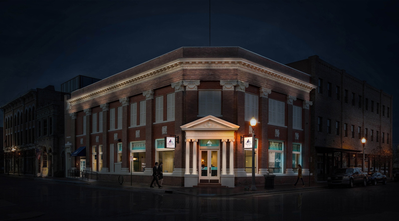 Hero image. Image of a large bank lit with lights in the front at nighttime.