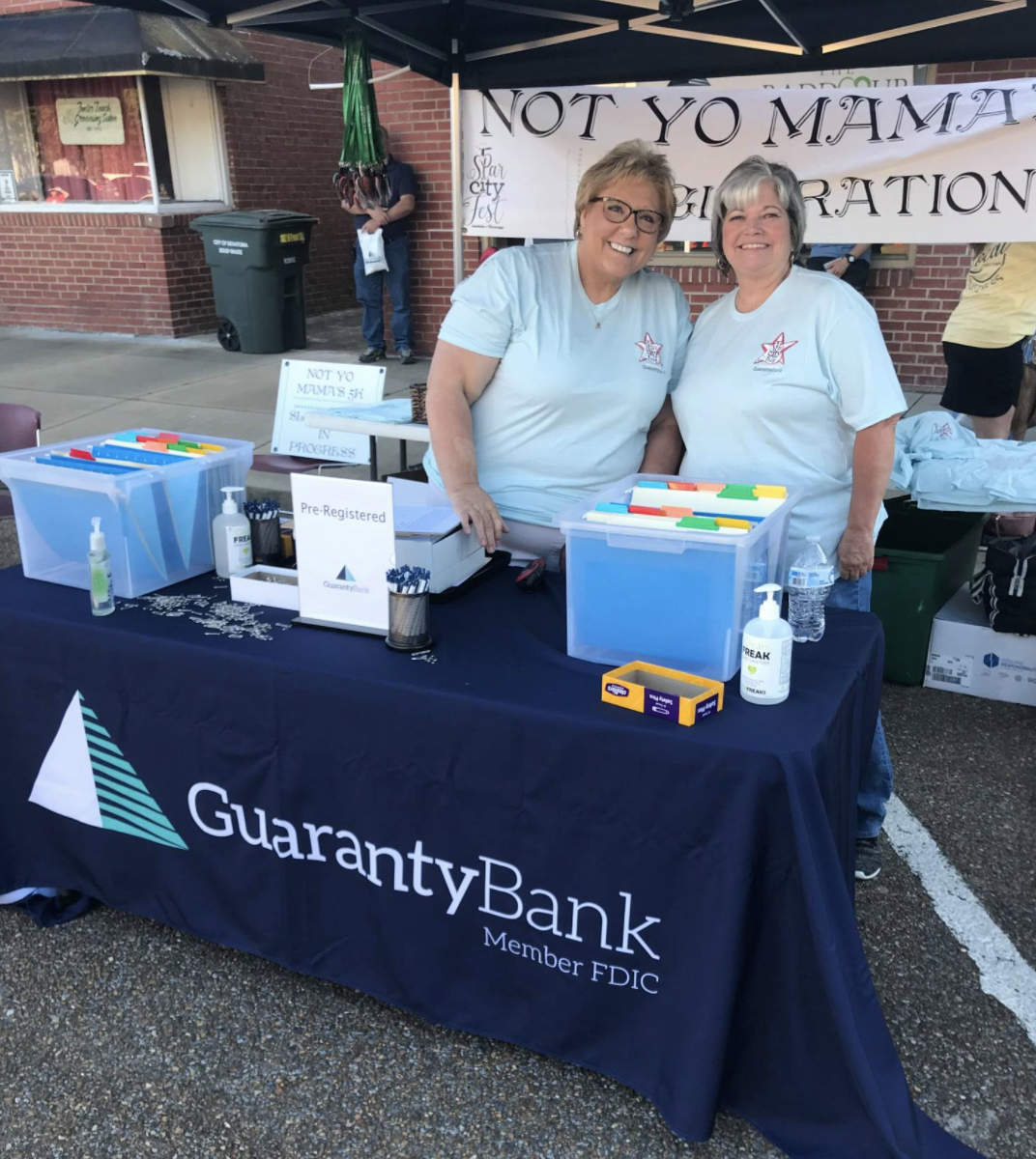 Two women behind a table and underneath a tent at a volunteering event. The two women are smiling as they stand behind the table with a blue table cover with the Guaranty Bank logo and a banner behind them. The table is set with bins, pens, and hand sanitizer.