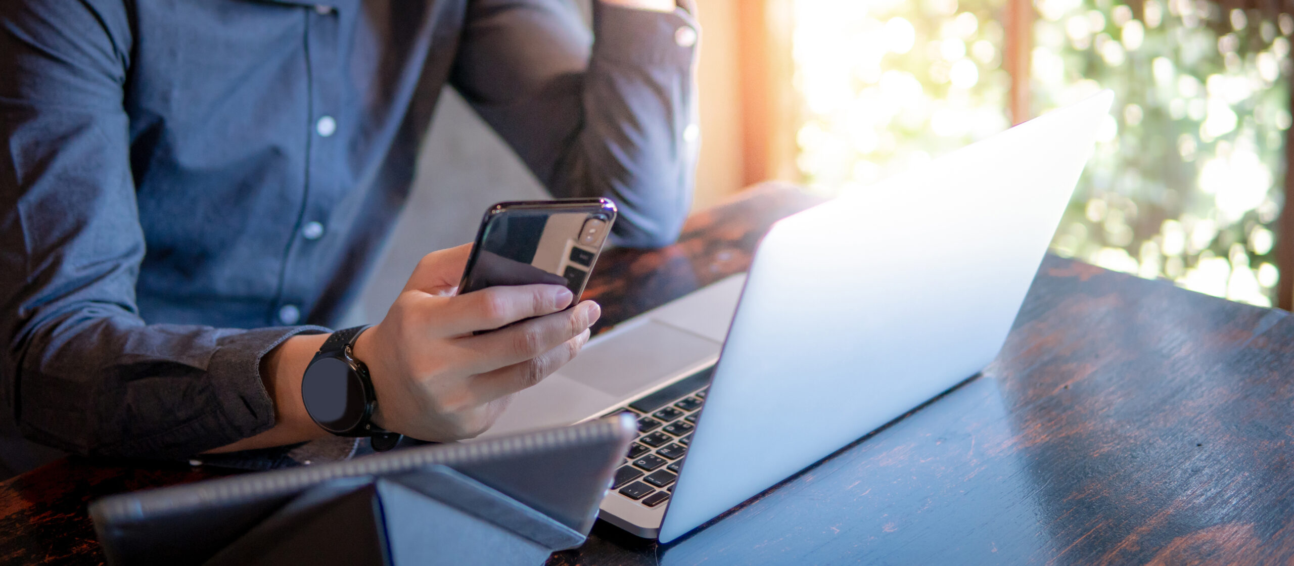 Male holding a smartphone sitting at a table with a laptop and digital tablet in front of him while working in a cafe.