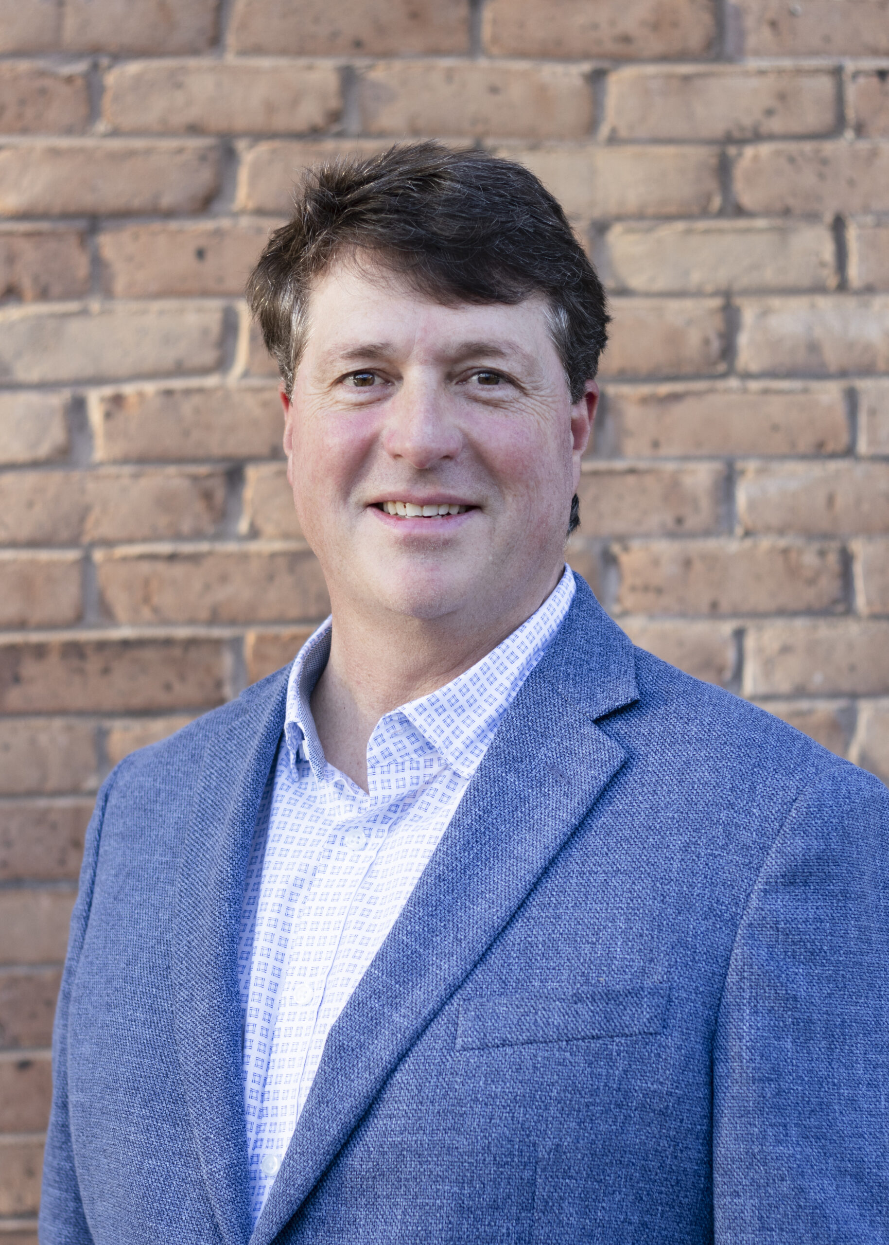 Headshot of Ward Gilbert. Man in blue suit jacket against a brick wall.